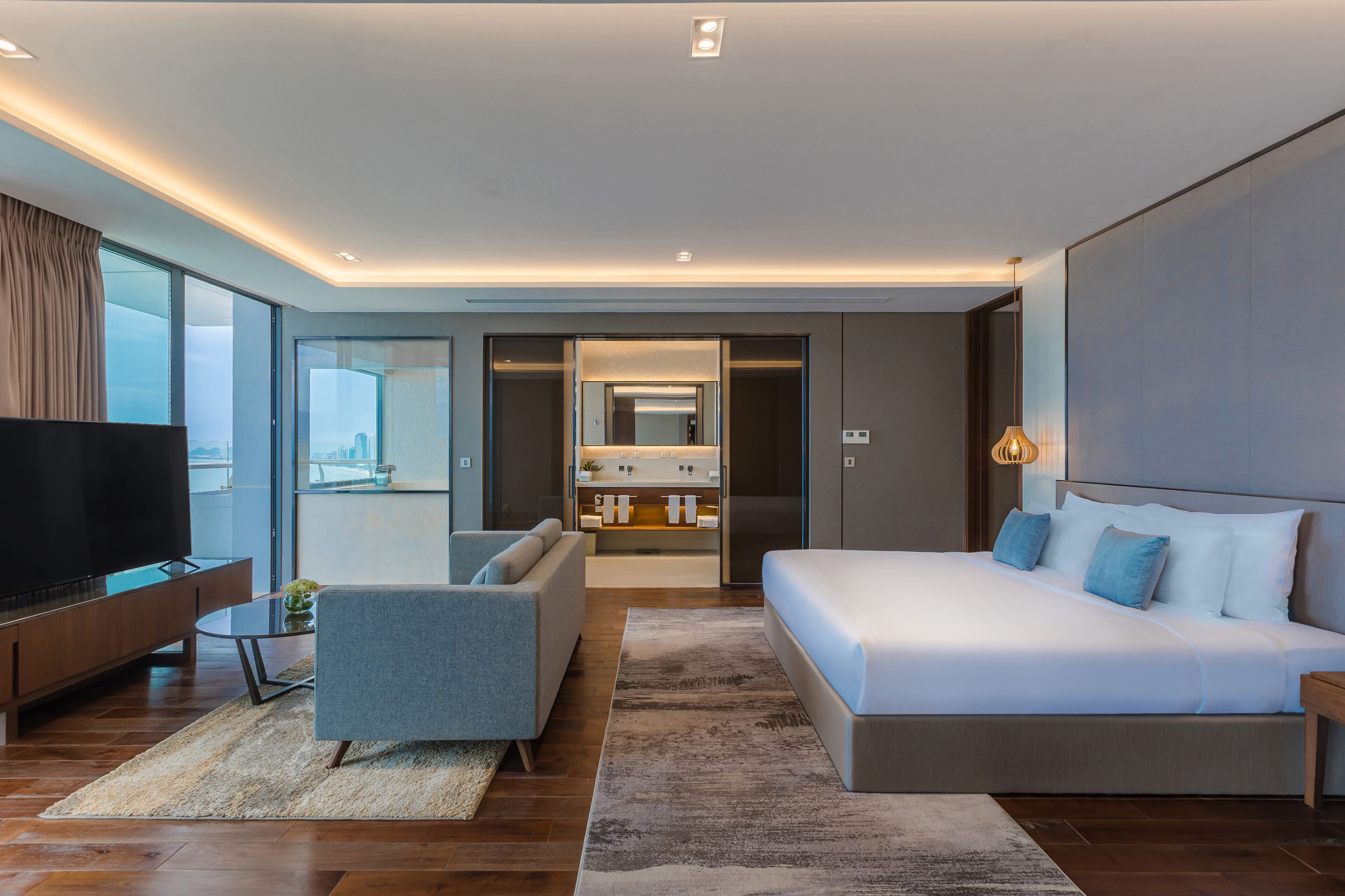 Habitación de hotel moderna con una cama grande, un sofá, un televisor y vistas de la ciudad a través de amplias ventanas.