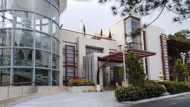 Un edificio moderno con fachada de vidrio, acceso cubierto y un área ajardinada con flores y arbustos.