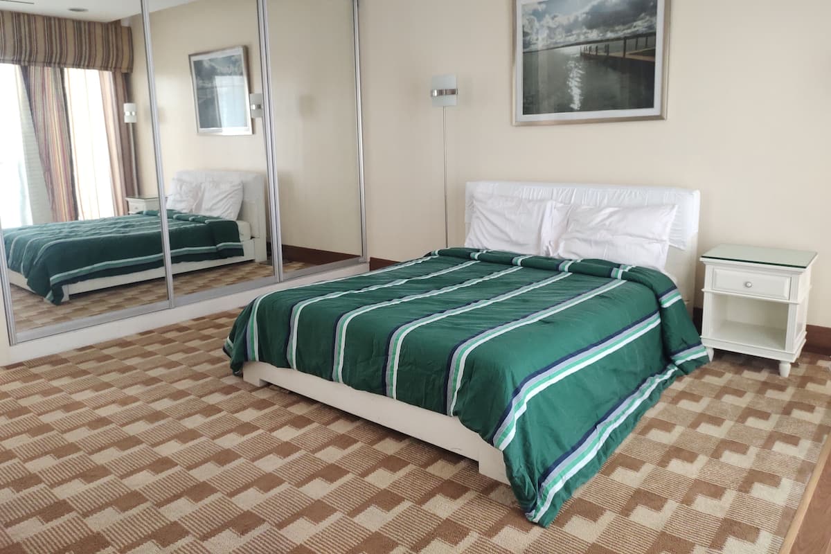 A bedroom with a large mirror, a bed with green bedding, a white headboard, a nightstand, and a painting on the wall.
