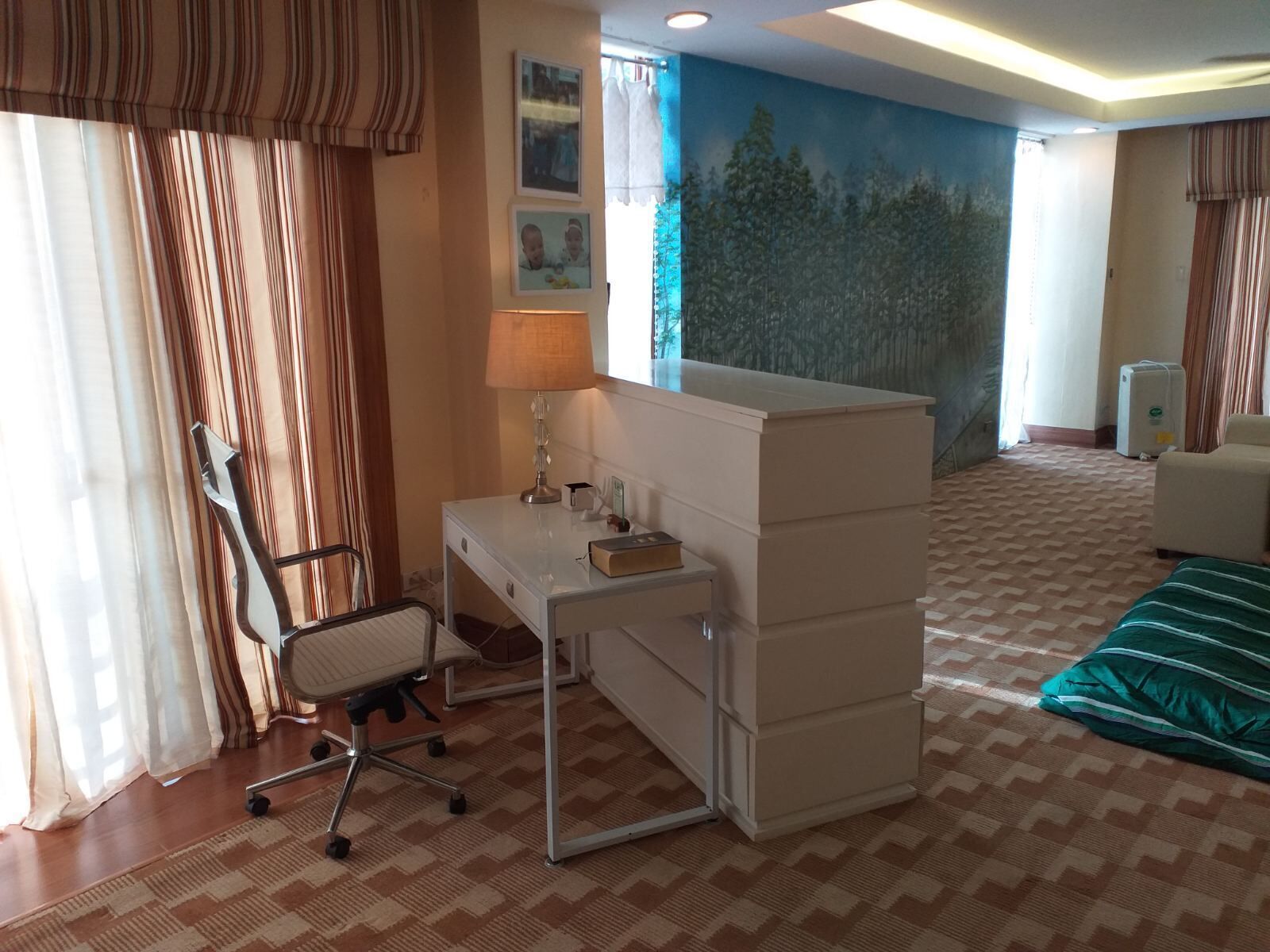 A hotel room with a desk, chair, and a bed. There is a window with curtains, a wall with a mural, and a radiator.