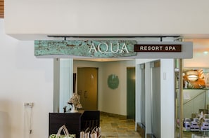 A sign for 'AQUA RESORT SPA' with a rustic design, a reception desk with a lamp, and a staircase leading to the spa area.