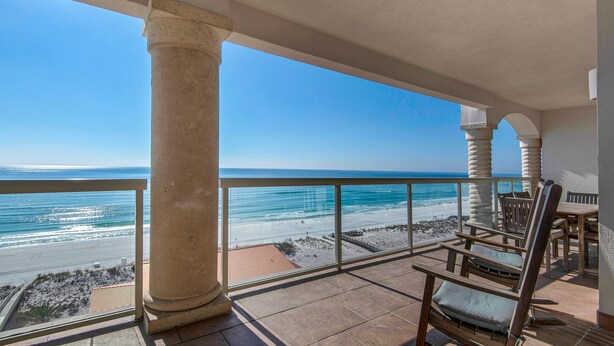 A balcony with a view of the beach, wooden chairs, and a table.