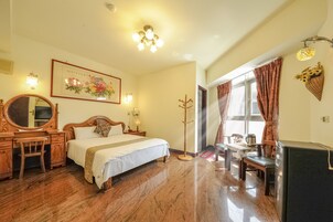 Un dormitorio bien iluminado con una cama grande, un aparador de madera, un escritorio y una silla. Se ve una ventana con cortinas, una lámpara de pared y un cuadro enmarcado sobre la cama.