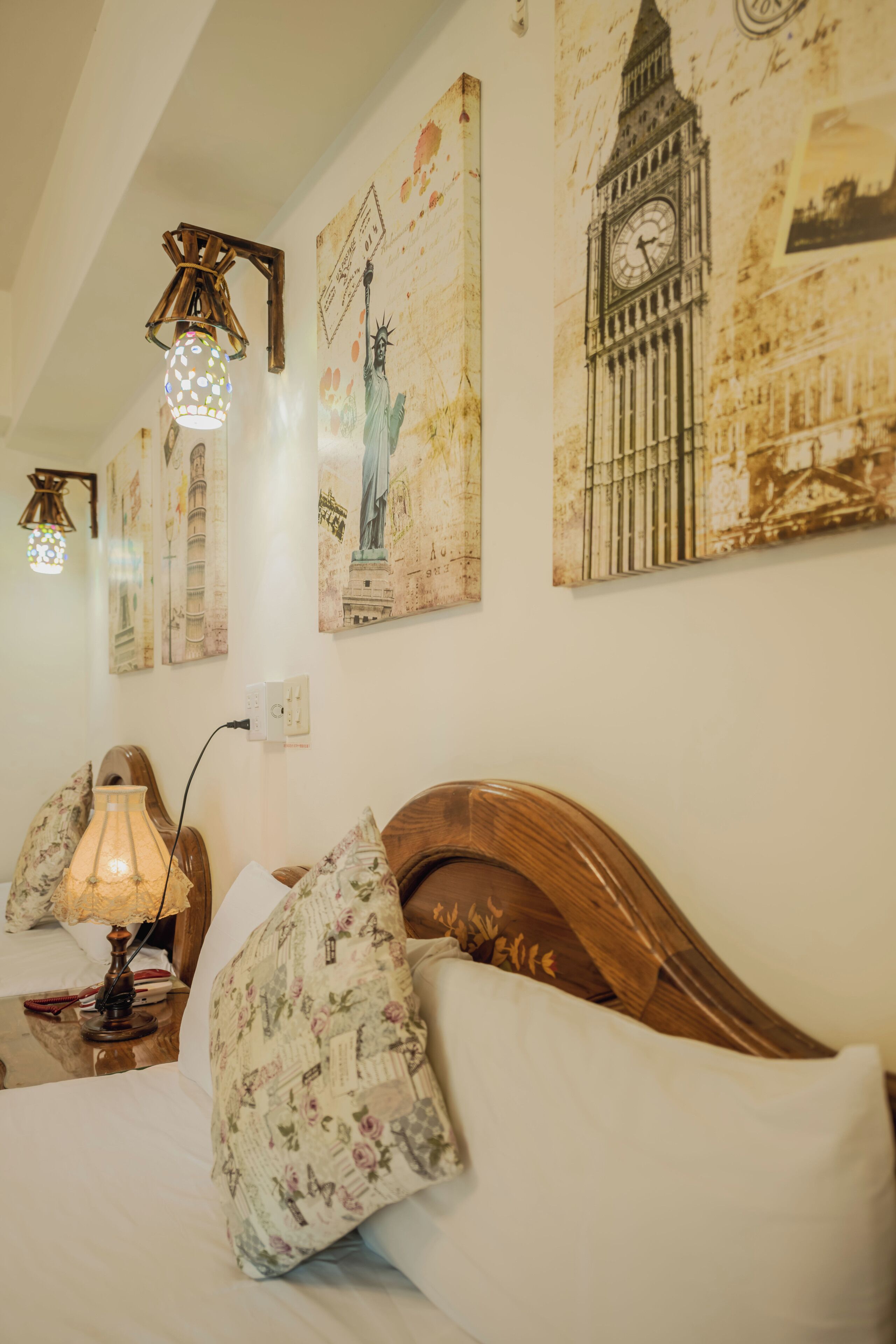 A bedroom with a wooden headboard, floral-patterned pillows, a lamp, and wall art depicting famous landmarks.
