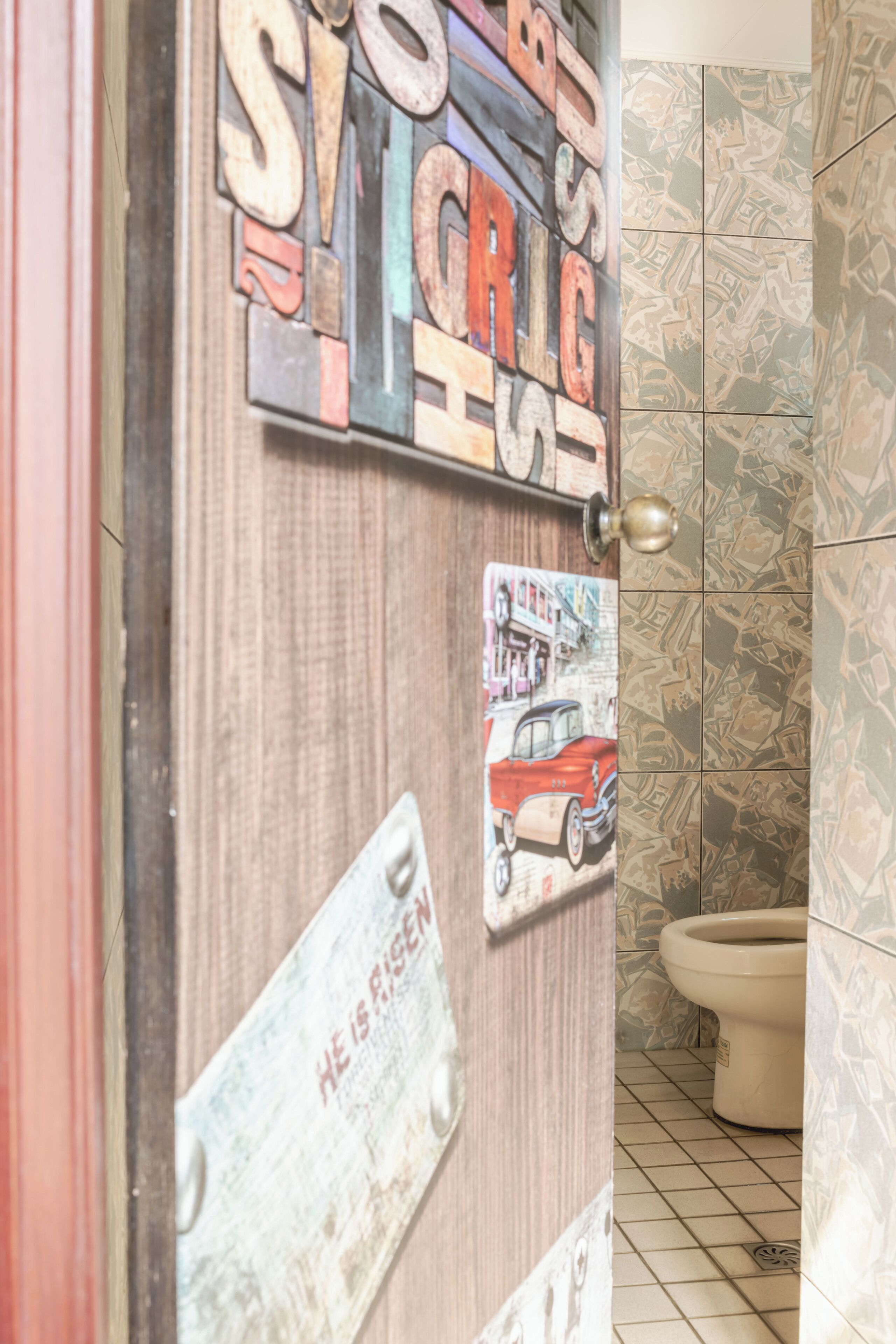 A bathroom with a toilet, a door with various stickers, and a patterned wall.