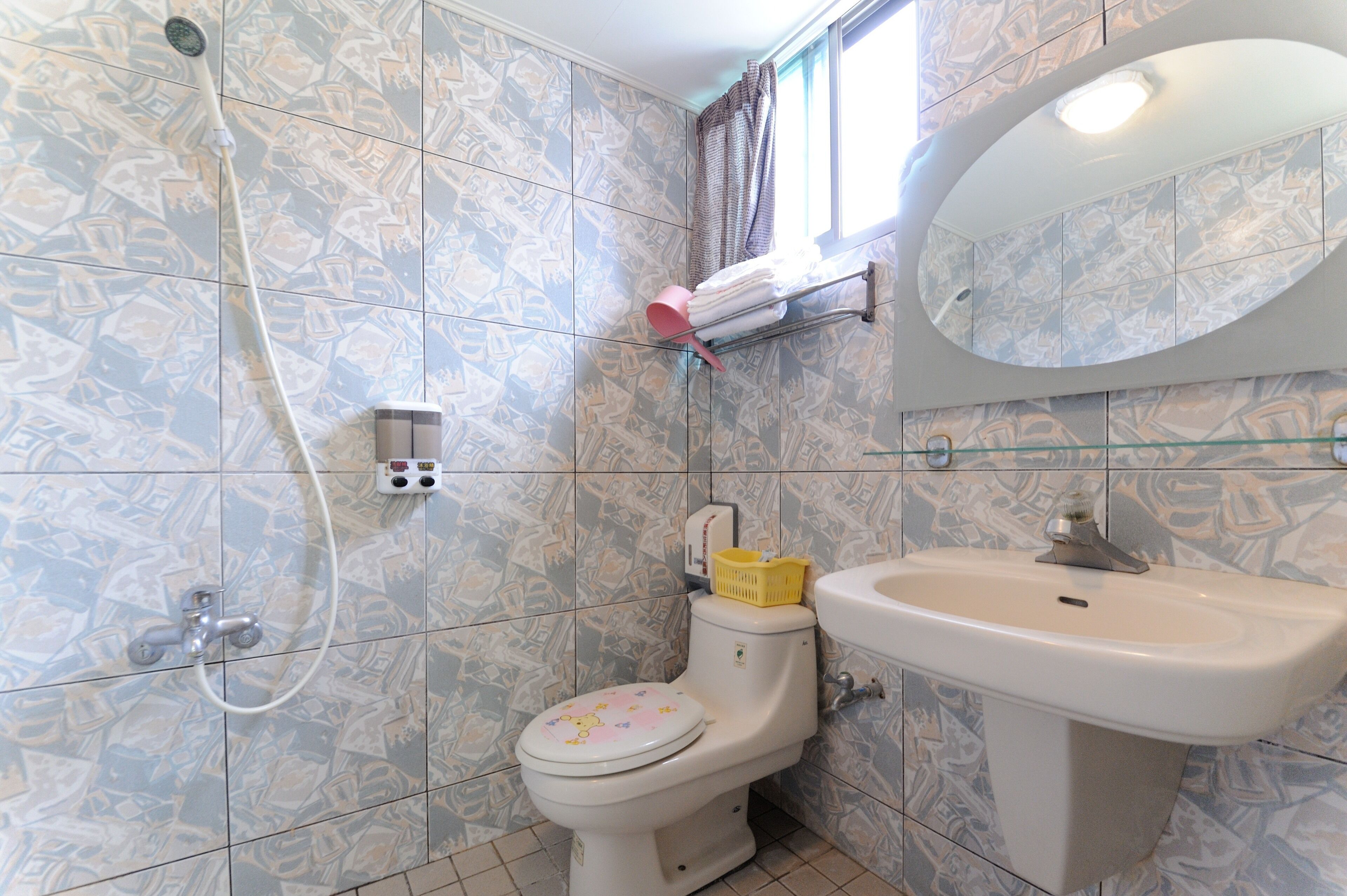 A bathroom with patterned tiles, a toilet with a pink seat cover, a sink, and a shower with a handheld showerhead.