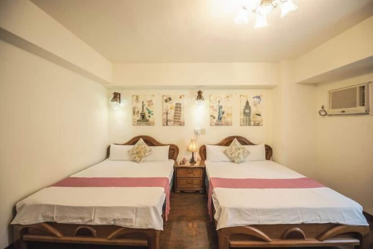A hotel room with two beds, a nightstand, a lamp, and wall art depicting famous landmarks.