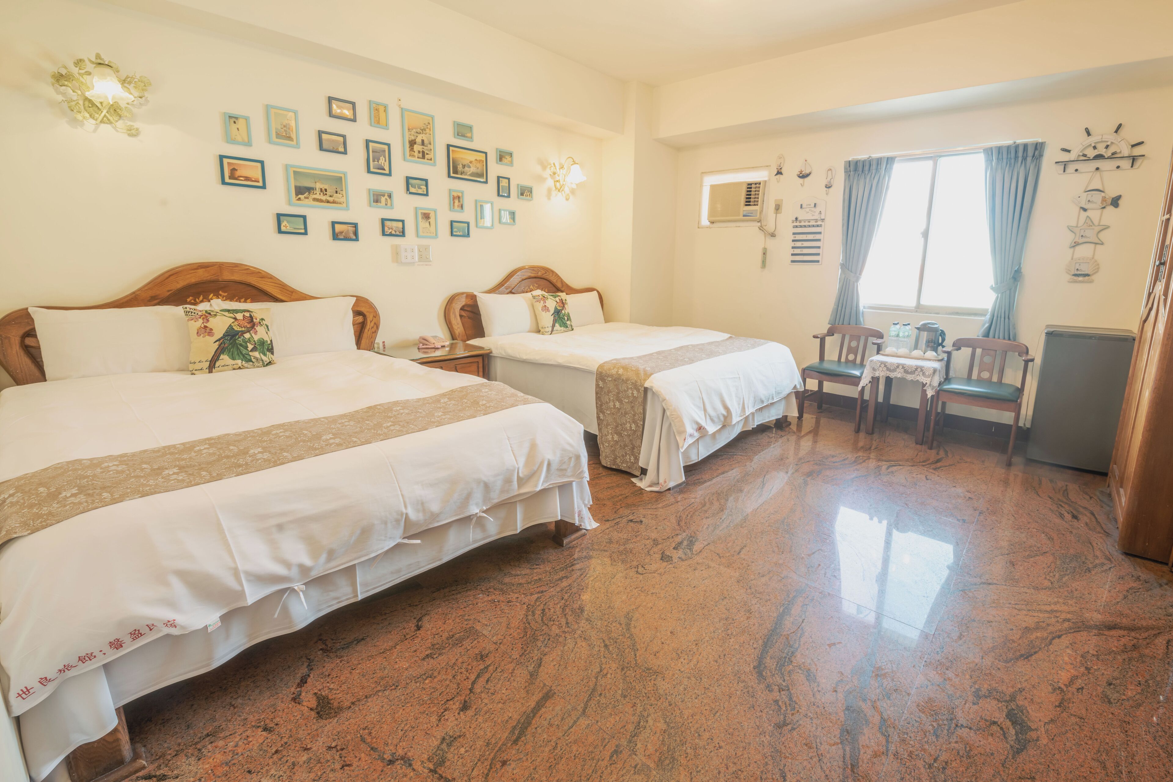 A hotel room with two beds, a desk, chairs, a window with curtains, and wall decorations.