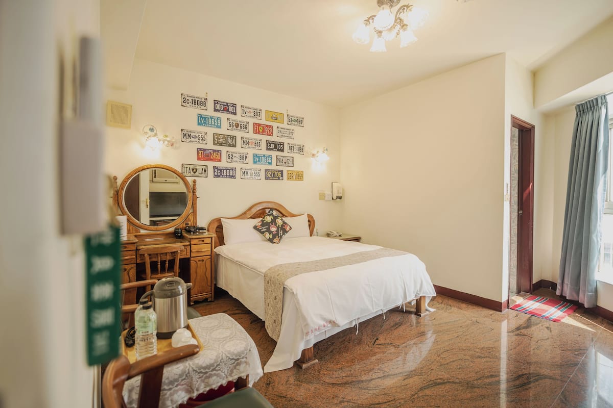 A hotel room with a bed, wooden nightstand, mirror, and wall-mounted license plates.