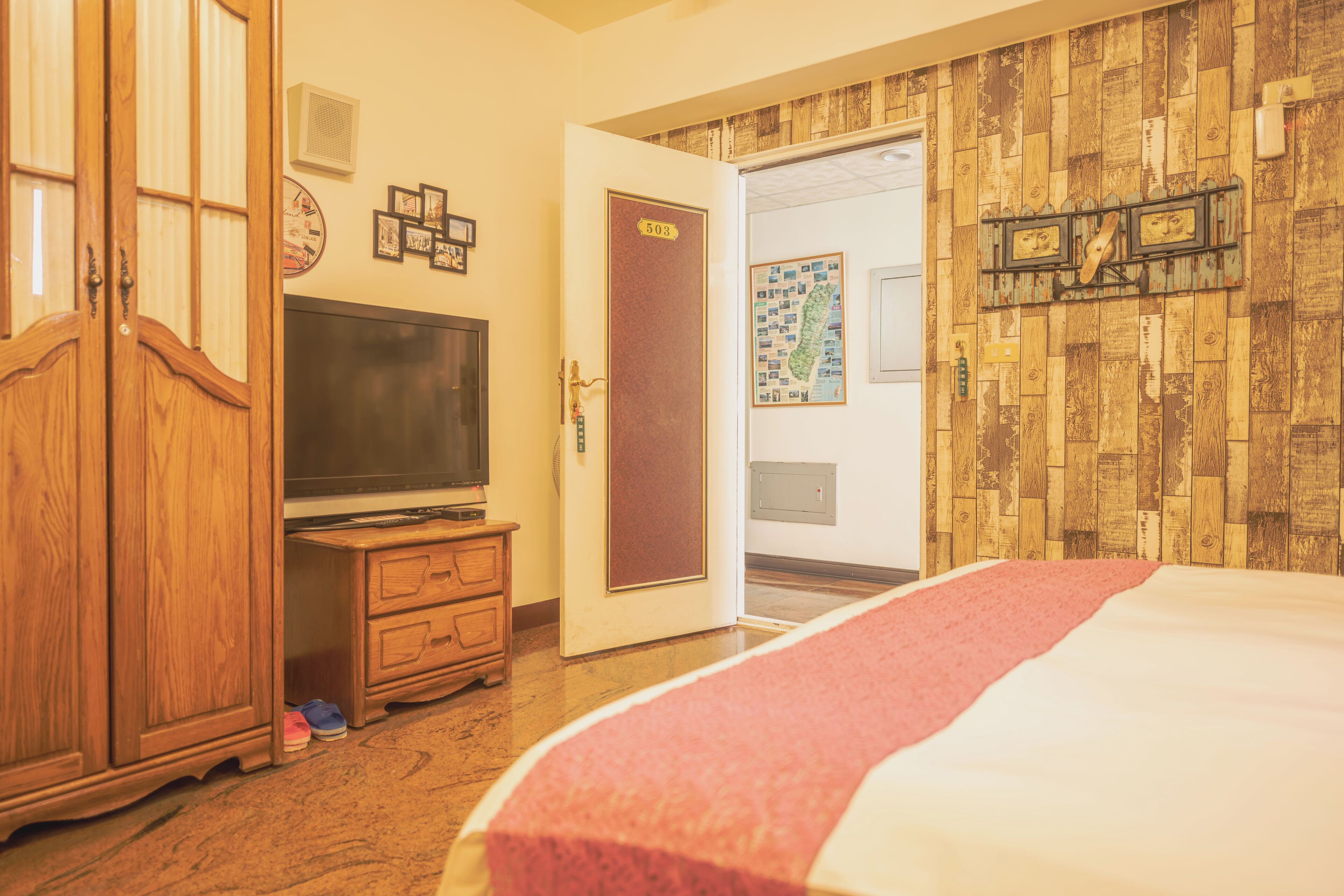 A room with a bed, a wooden wardrobe, a television on a stand, and a door with a number sign.