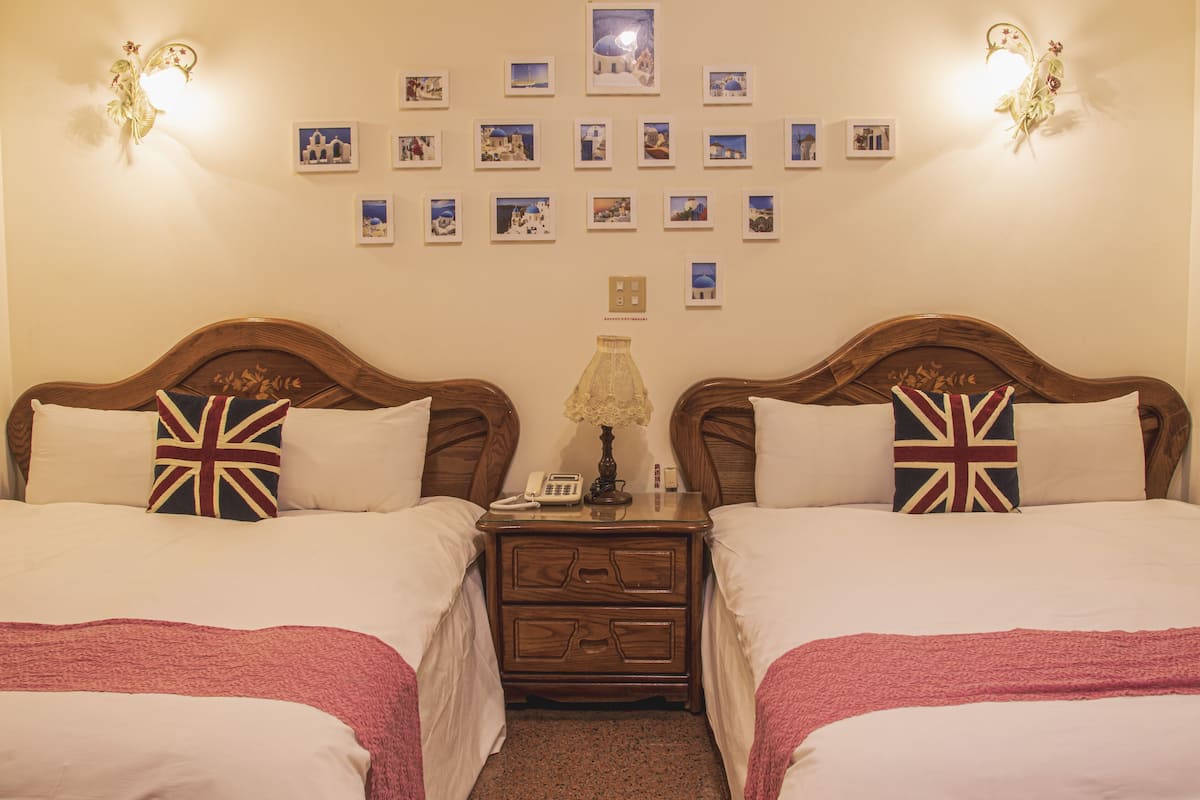 A double room with two beds, each with a pillow featuring the Union Jack design. There is a nightstand between the beds with a lamp and a phone. The wall above the beds is decorated with framed photographs.