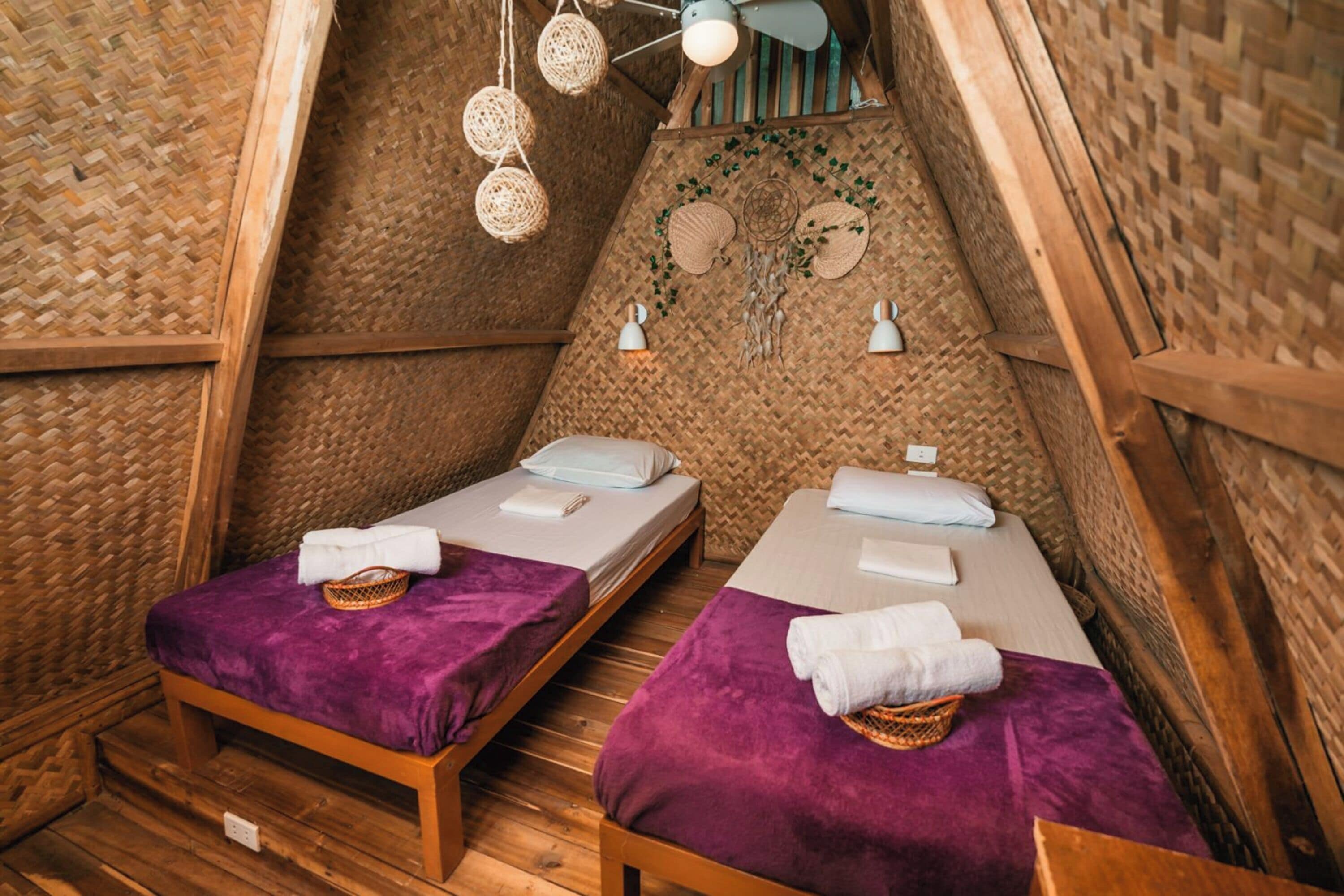 A cozy, rustic room with two wooden beds, woven walls, and a ceiling fan.