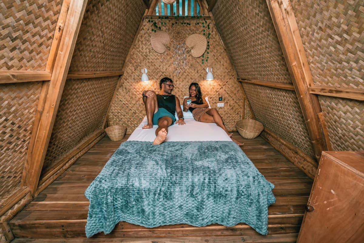 A cozy, triangular room with a woven ceiling and walls, a bed with a blue blanket, and two wall-mounted lamps.