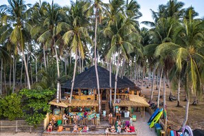 A beachside resort with multiple wooden structures, including a bar area with outdoor seating and a staircase leading to an upper level.