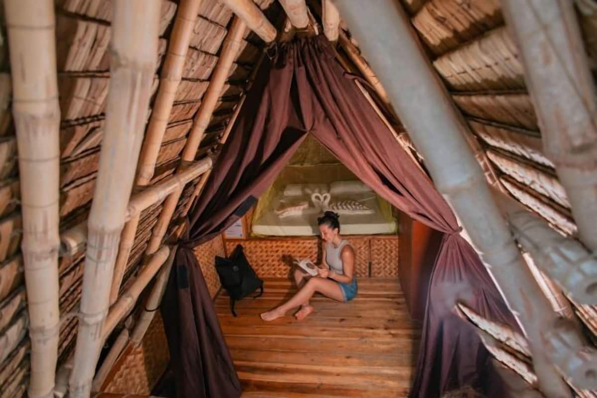 A person sitting in a bamboo hut with a bed and a small table.