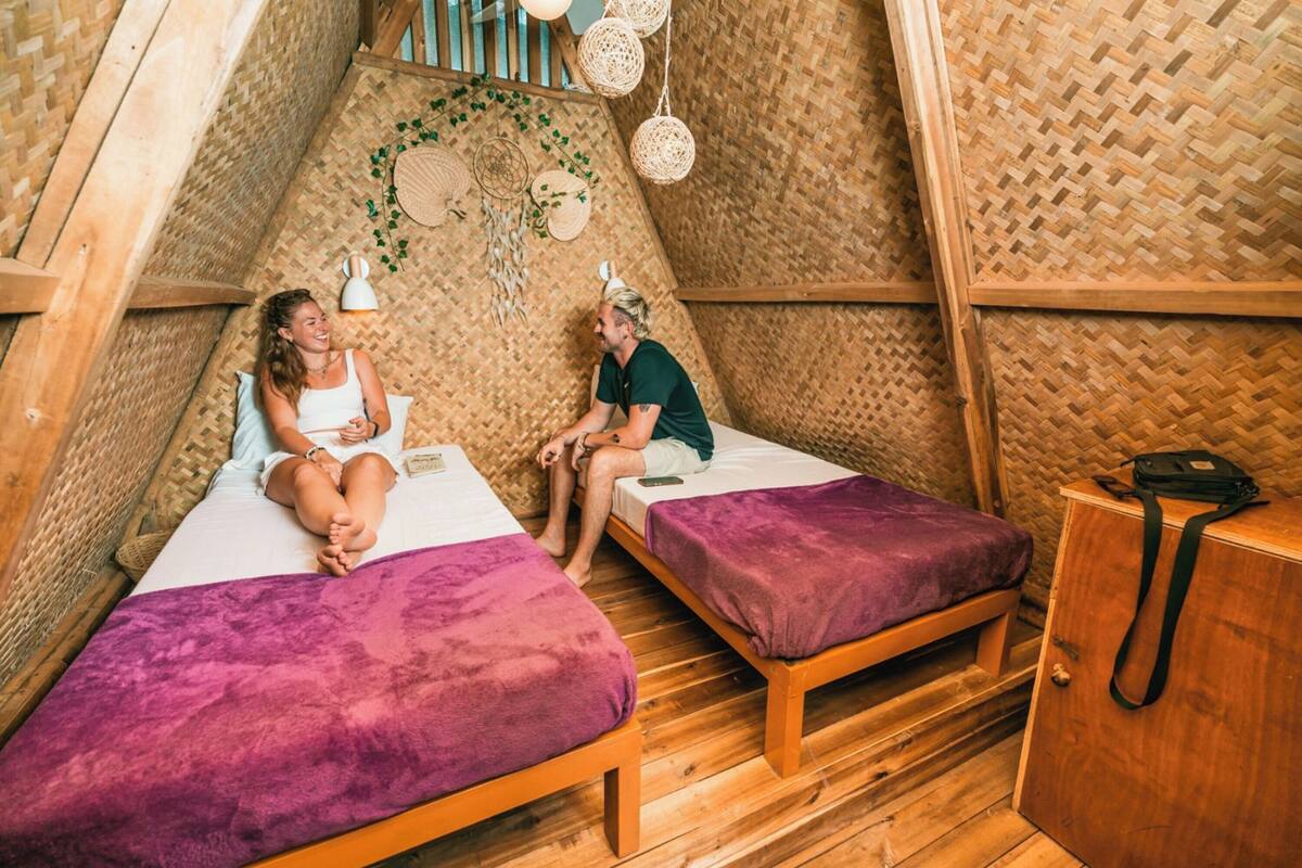 A cozy, triangular room with two beds, each with a purple blanket. The walls are made of woven material, and there are woven hanging lights.