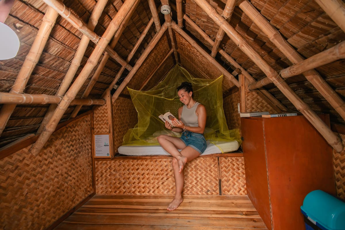 A person sitting on a bed in a room with a thatched roof and wooden walls.