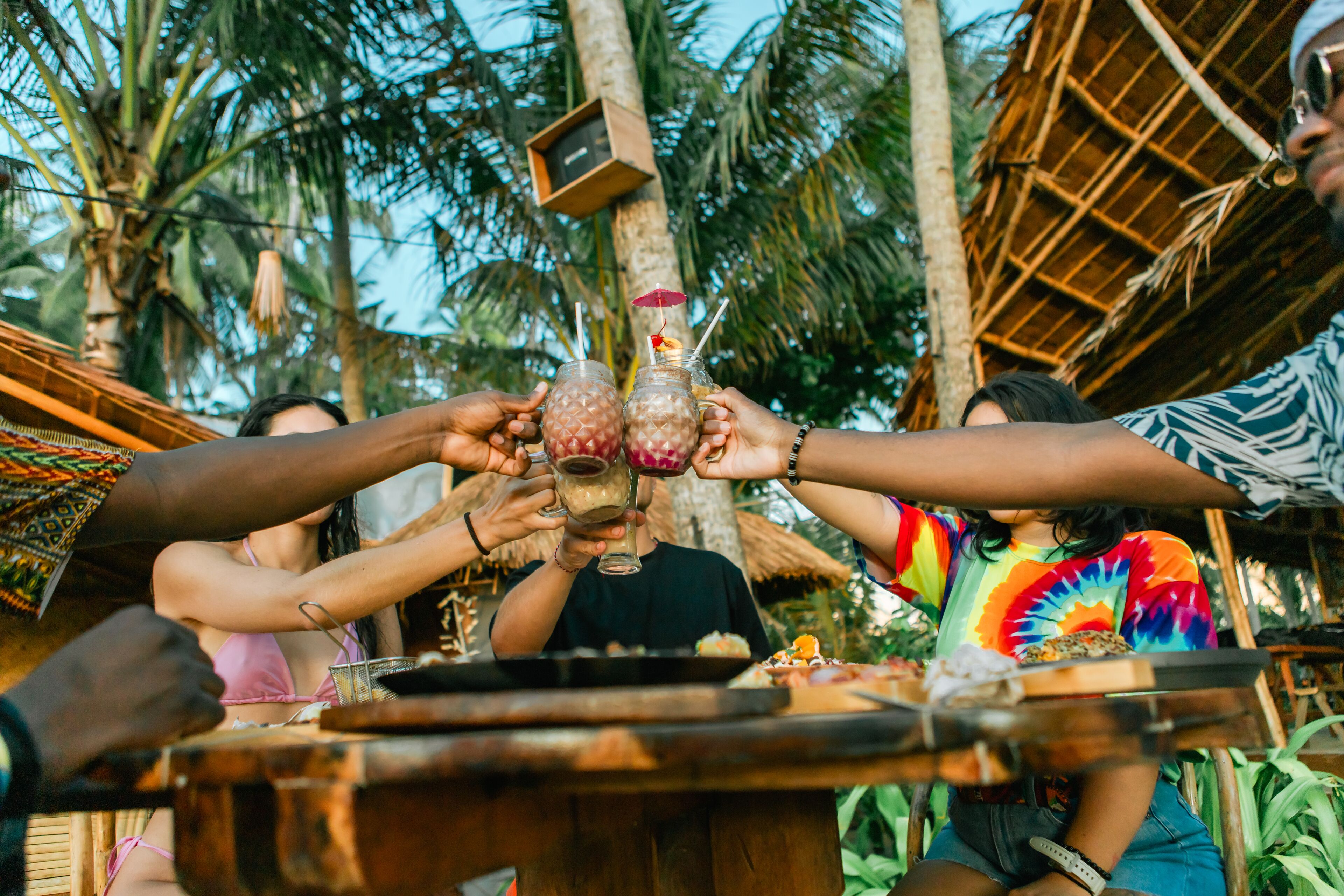 Un groupe de personnes trinquant avec des boissons autour d’une table en bois, à l’extérieur, entourés de palmiers et d’une structure à toit de chaume.
