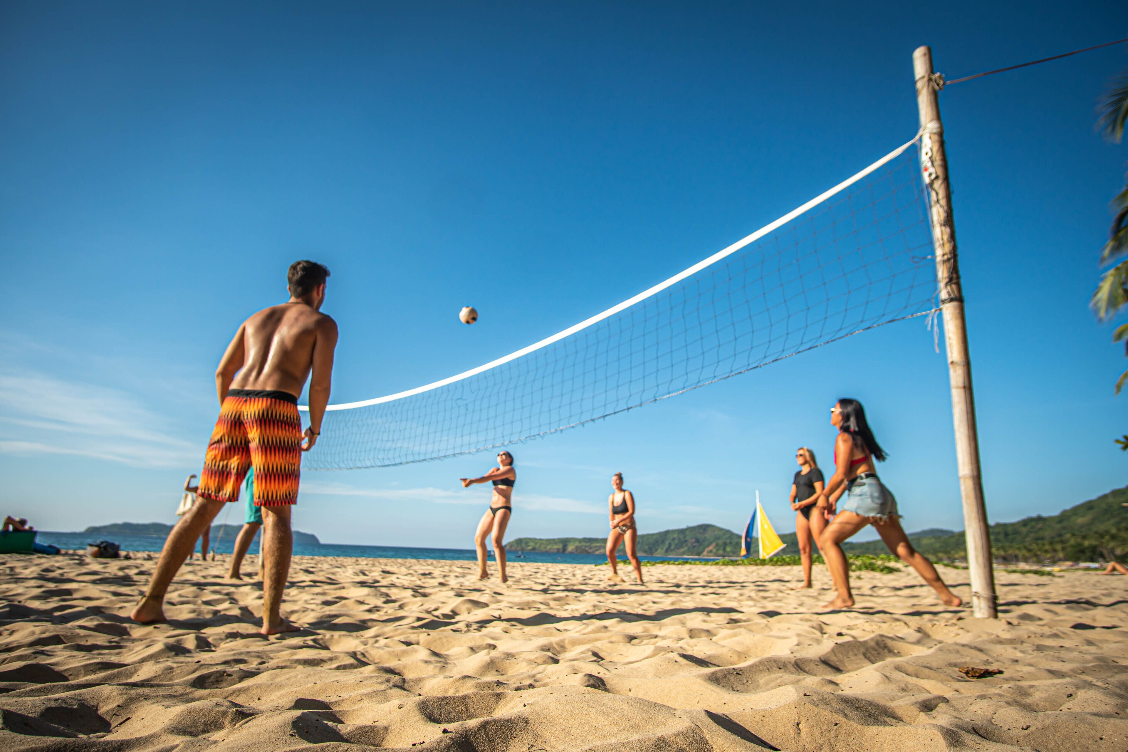 A beach volleyball game with players in swimwear, a volleyball net, and a clear sky.