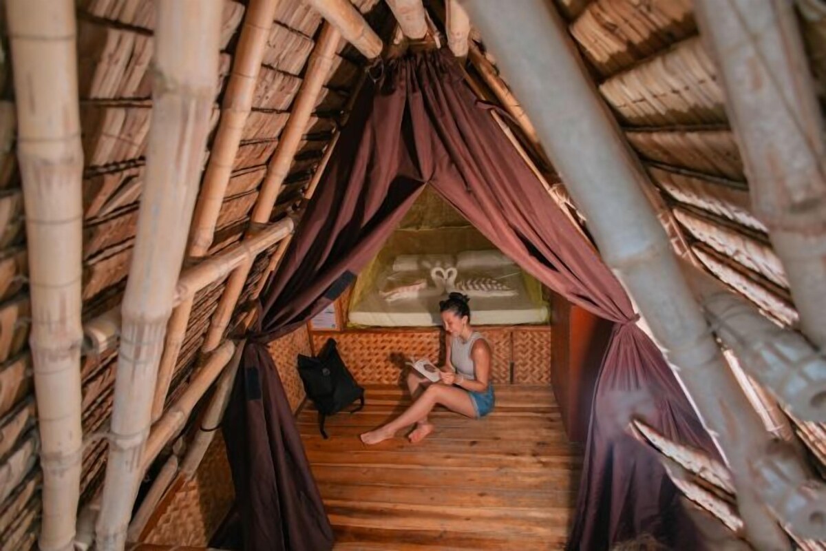 A person sitting in a bamboo hut with a bed and a small table.