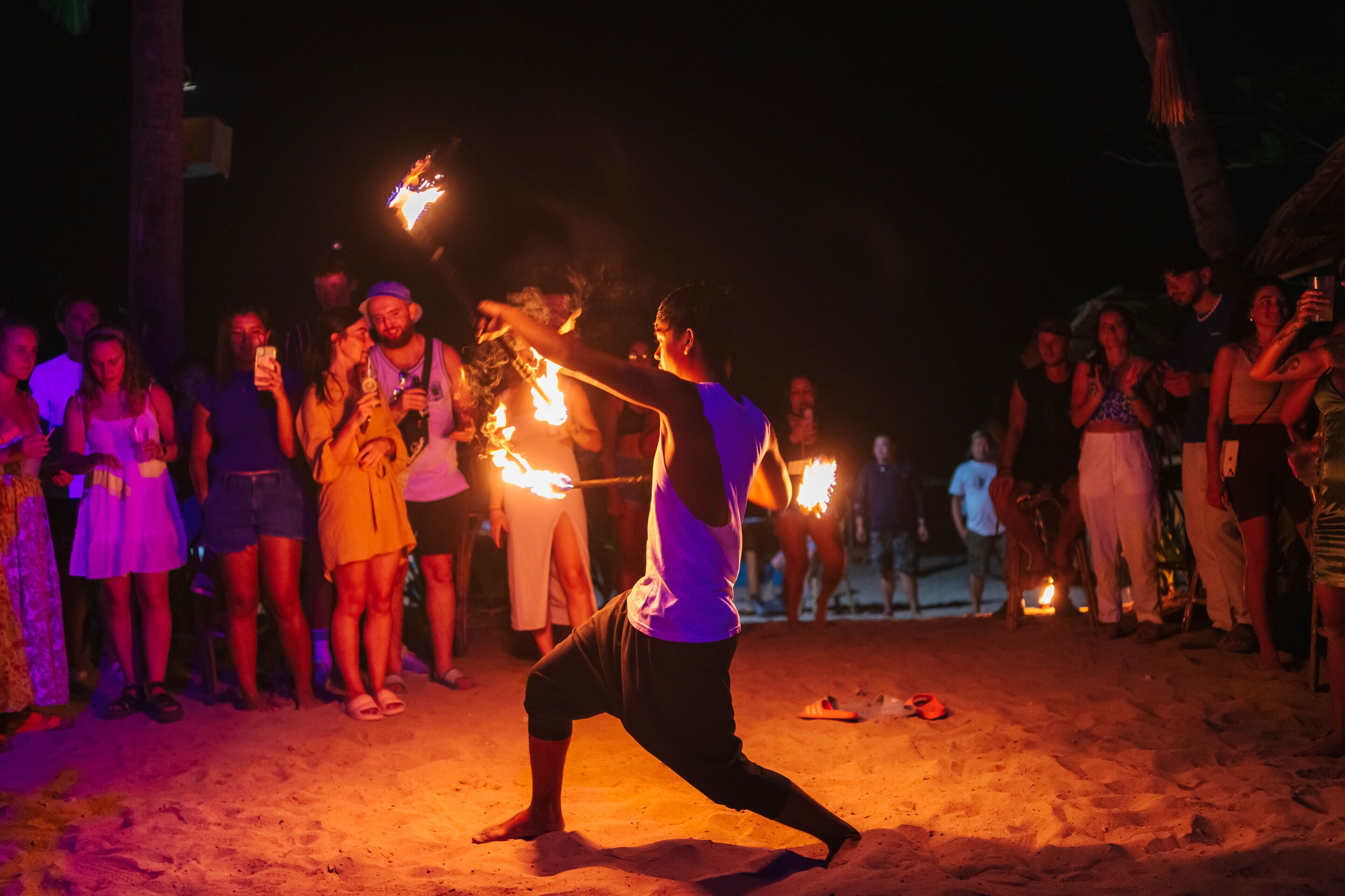 A person performing with fire props in a sandy outdoor setting, surrounded by an audience.