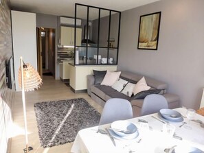 A modern living room with a grey sofa, a dining table set with plates and glasses, a black-framed mirror, and a kitchen in the background.