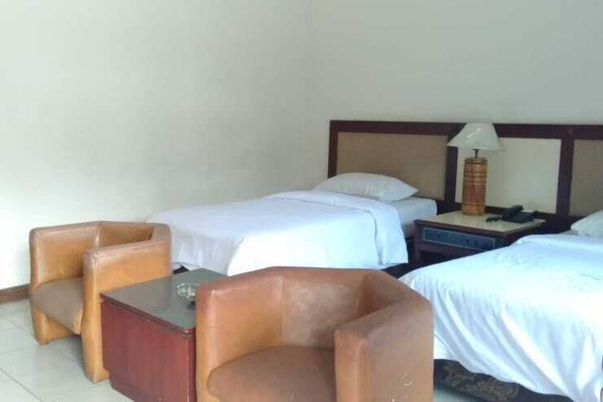 A hotel room with two beds, a wooden nightstand, a lamp, and two leather armchairs.