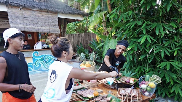 A wooden table with skewers of meat and vegetables, a basket of fruits, and a drink in a glass.