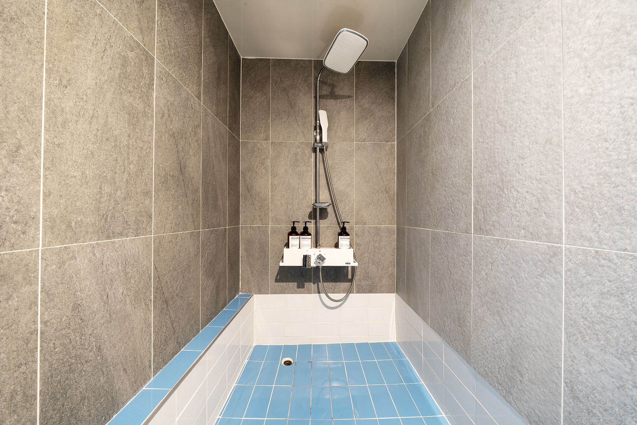 A shower with a handheld showerhead, a handheld soap dispenser, and a bottle of shampoo.