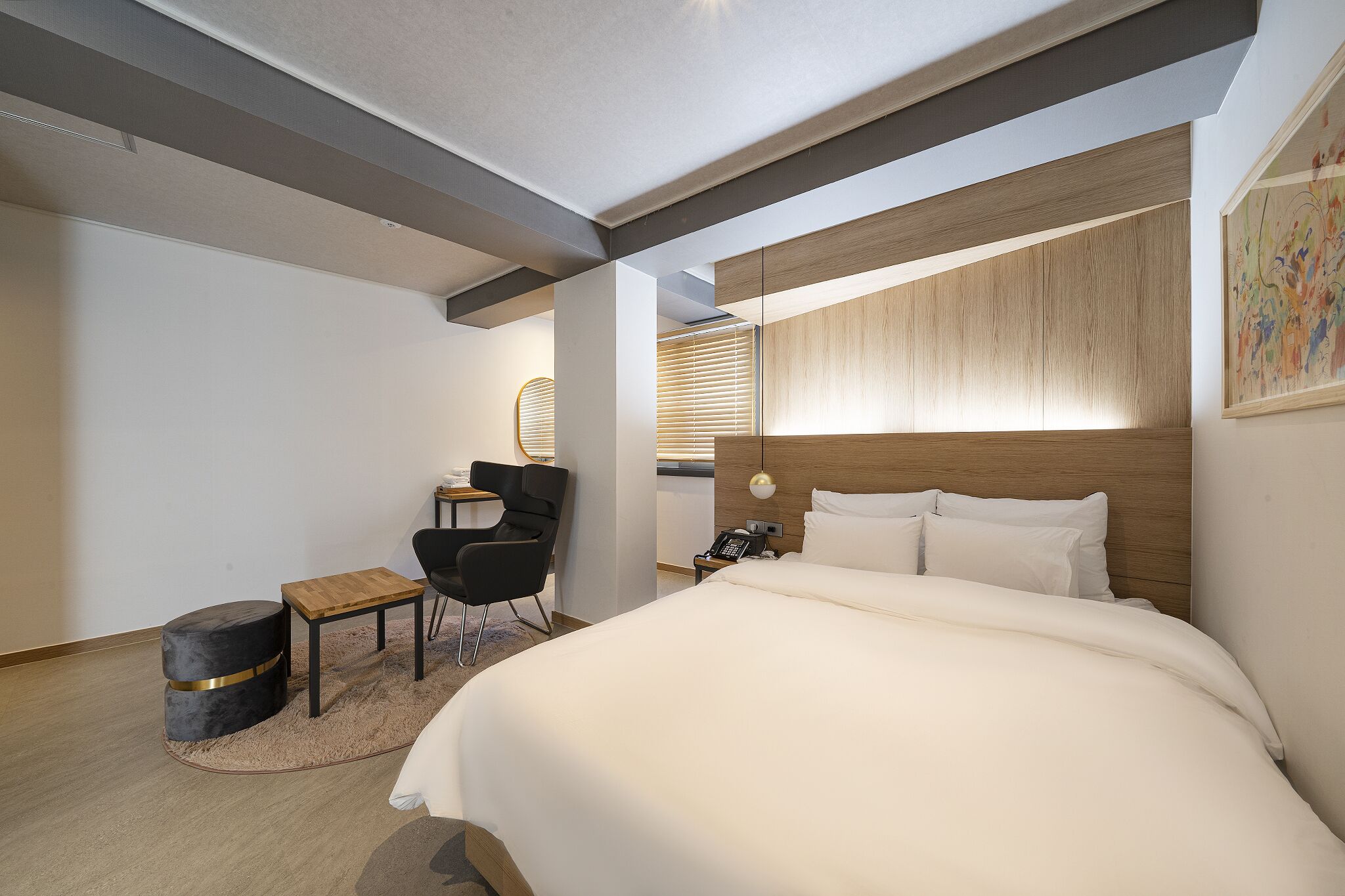 A modern hotel room with a large bed, a desk, a chair, and a small round ottoman.