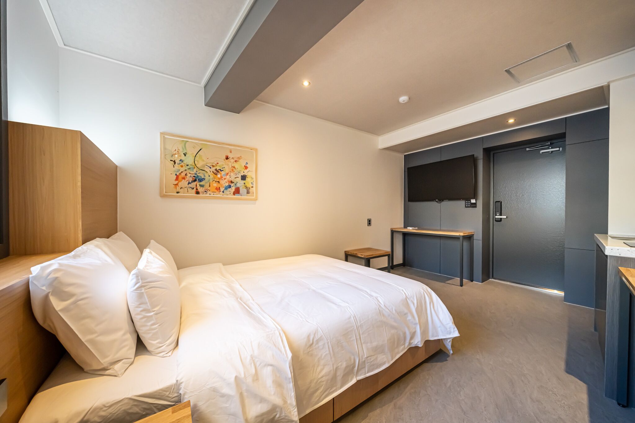Un dormitorio moderno con una cama grande, un cabecero de madera, una mesita de noche pequeña, un escritorio y una puerta que da a un baño.