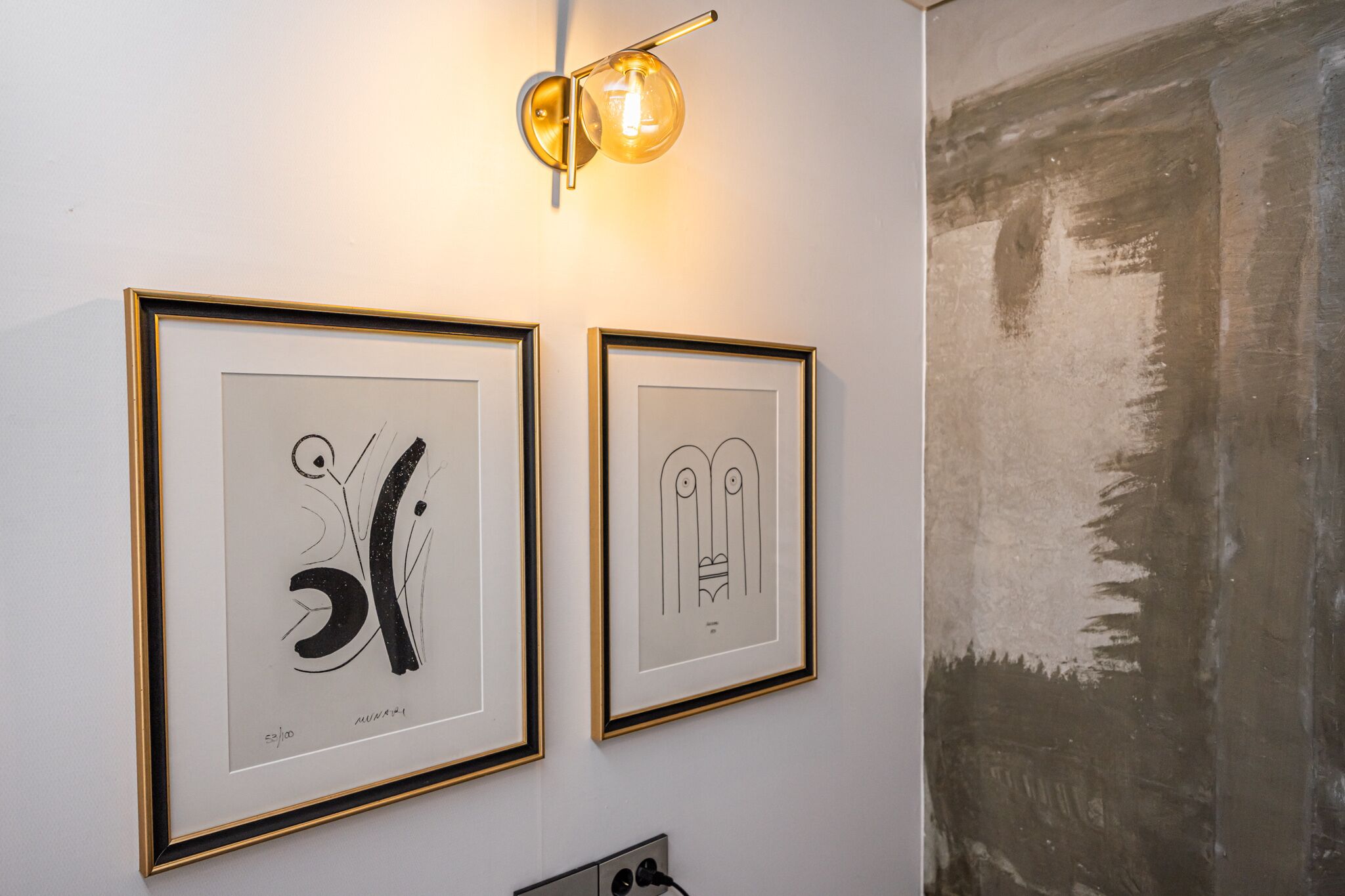 Two framed abstract artworks on a wall, a wall-mounted light fixture, and a textured wall to the right.
