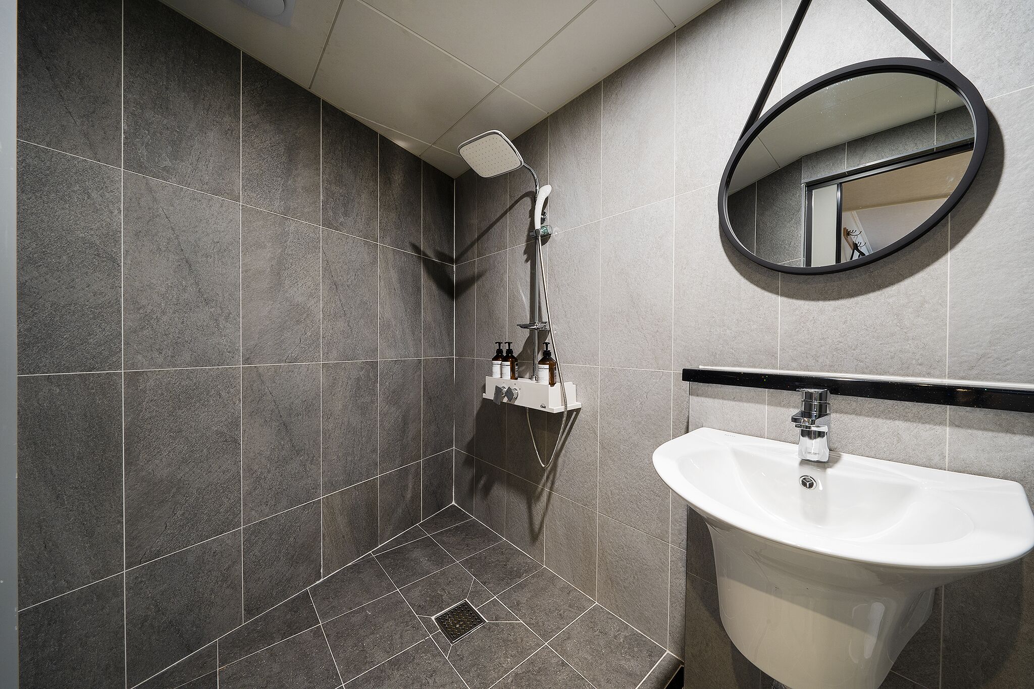 Een moderne badkamer met grijze tegels, een douche, een wastafel en een spiegel.