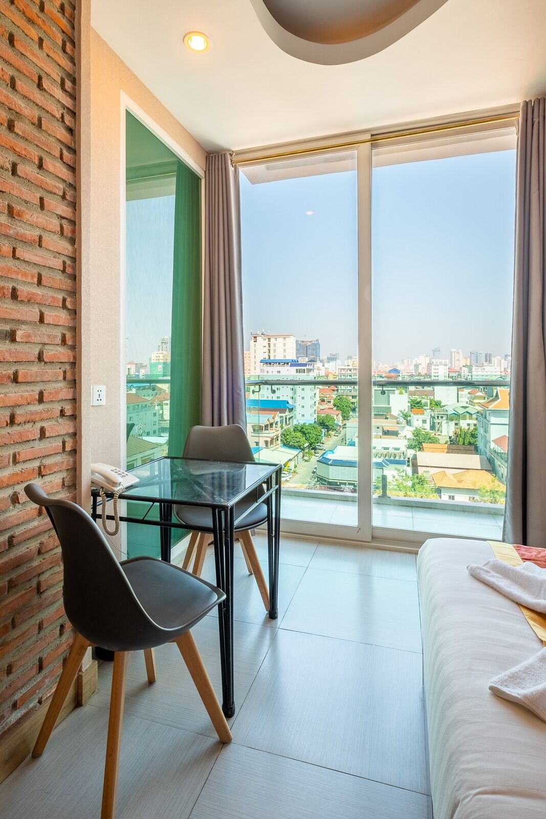 A modern hotel room with a glass table, two chairs, a bed, and a city view.