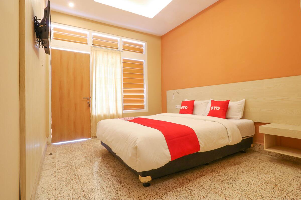 A bedroom with a bed, a bedside table, a window with blinds, and a door.