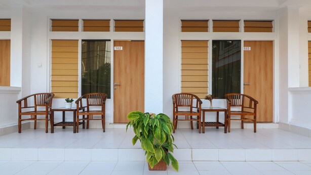 Un hall d’entrée propre et moderne, avec deux portes en bois portant les numéros 102 et 103, une plante en pot placée au centre, ainsi qu’un ensemble de chaises et de tables en bois disposées pour accueillir les visiteurs.