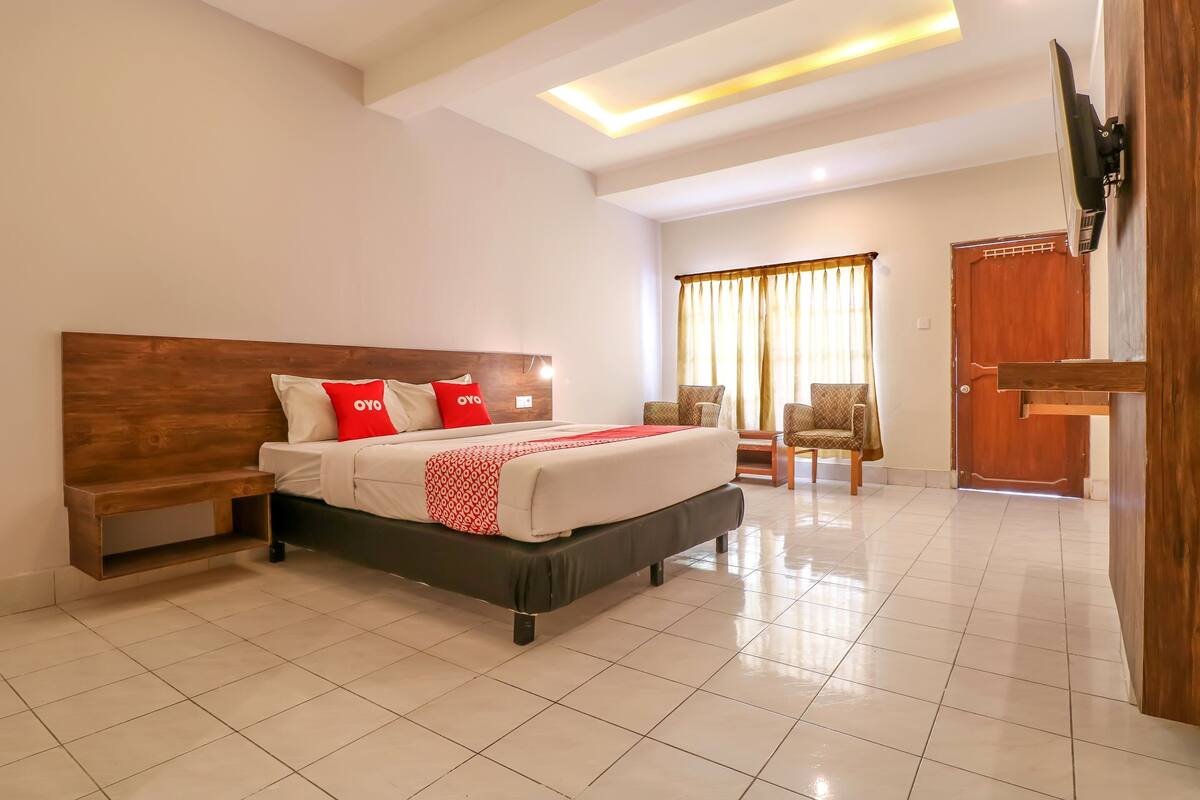 A hotel room with a large bed, a wooden headboard, a bench, a TV mounted on the wall, and a door.