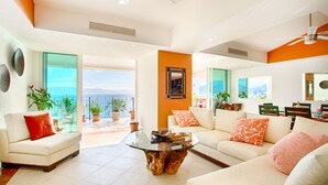 A spacious living room with a glass coffee table, two white sofas with orange and pink cushions, and a view of the ocean through large windows. - View and Breeze  w/cleaning service (Las Glorias)