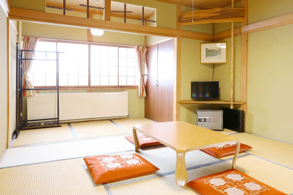 Une pièce de style japonais traditionnel, avec un sol recouvert de tatamis, une table basse, des coussins, un radiateur, un téléviseur et une étagère sur laquelle est posée une image.
