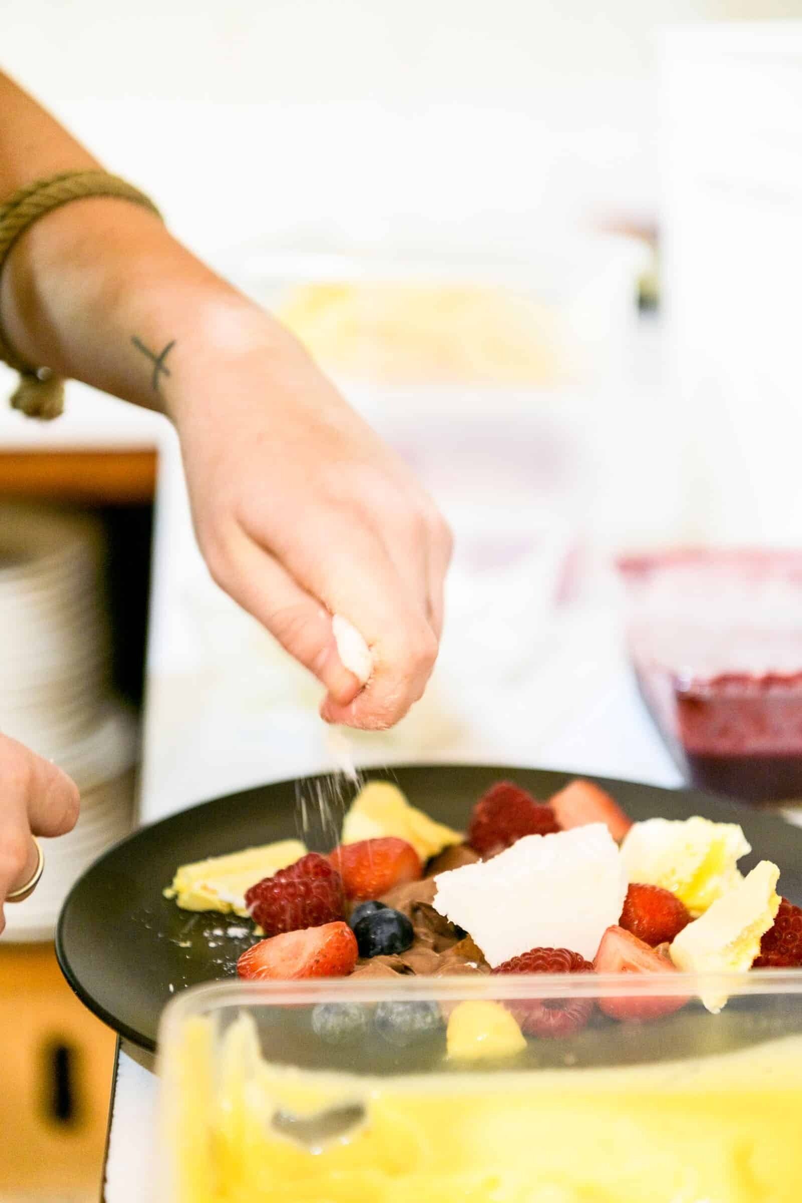 A person sprinkling powdered sugar over a dessert with strawberries and blueberries.