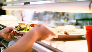 Una persona sosteniendo un plato con una ensalada y un sándwich en una mesa, con una ventana al fondo.