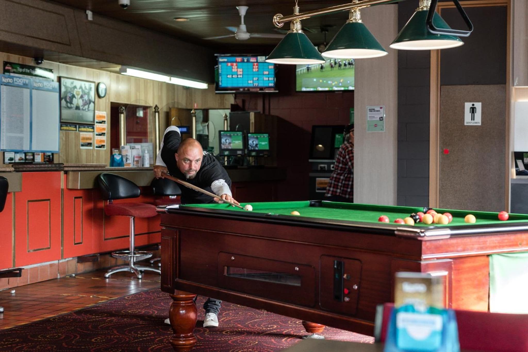 Una mesa de billar con una persona jugando, una zona de bar con taburetes y un televisor mostrando un partido deportivo.