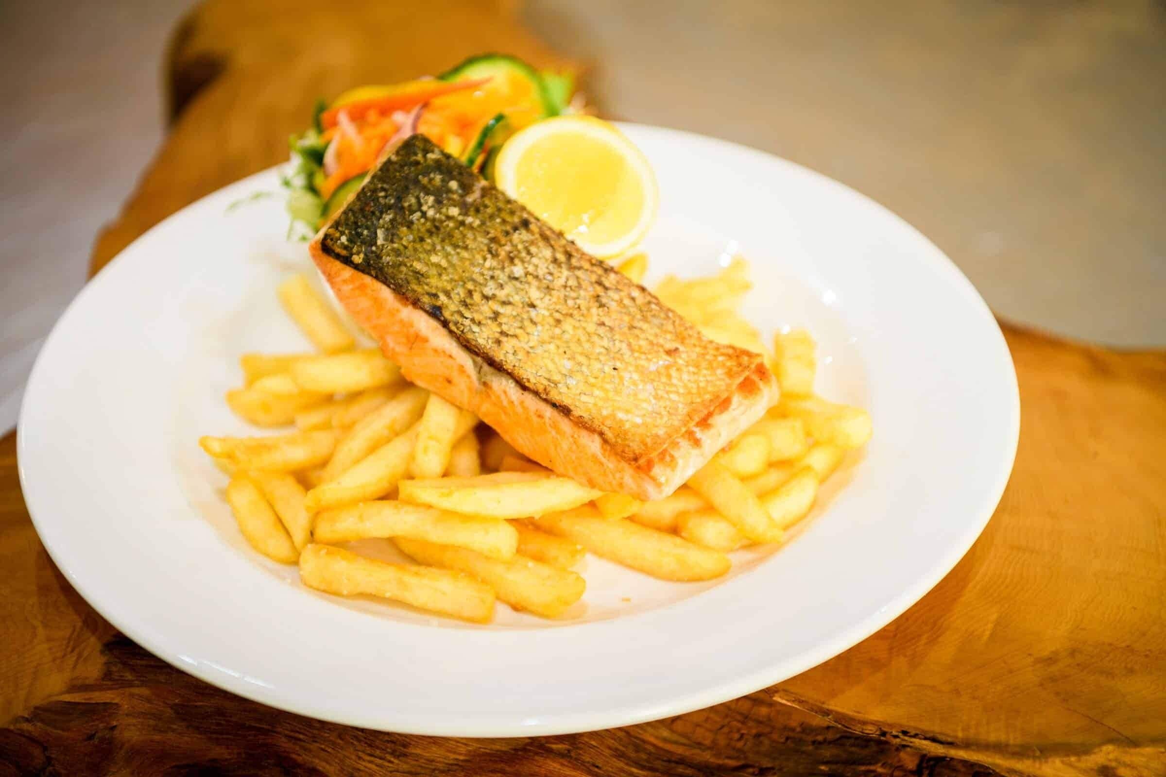 A plate with a piece of salmon, French fries, a slice of lemon, and a side salad.