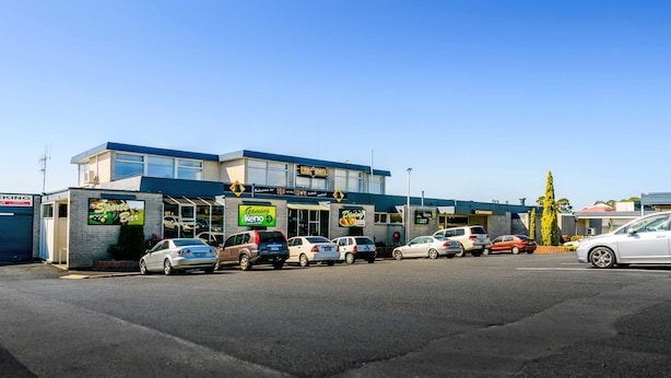 Un edificio comercial que alberga varios negocios, incluyendo un bar deportivo, una zona de juegos y una tienda de vidrio. Hay varios coches estacionados adelante.