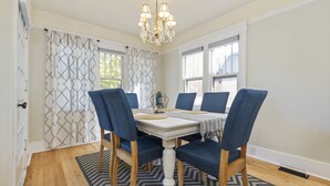 Comedor con mesa blanca, sillas azules, una araña y cortinas estampadas.