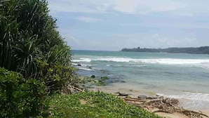 A coastal landscape with a sandy beach, dense green vegetation, and a view of the ocean with waves. - Palm Hill Guest House (Talalla)