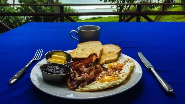 A breakfast plate with toast, eggs, and bacon, a mug, and a bowl of jam on a table with a blue tablecloth, overlooking a scenic view. - Palm Hill Guest House (Talalla)