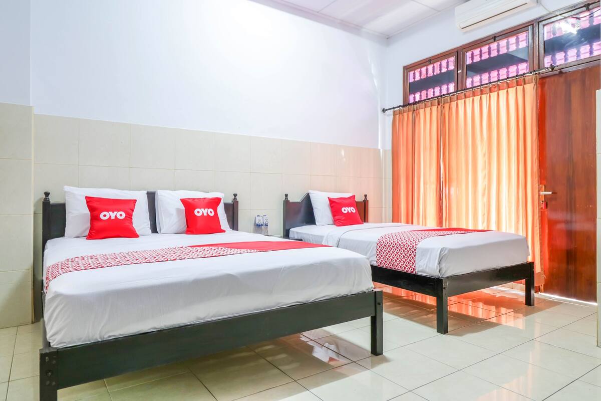 A hotel room with two beds, each with white bedding and red pillows, and a large window with orange curtains.
