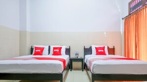 Une chambre d’hôtel avec deux lits, chacun doté de draps blancs et d’oreillers rouges, une petite table entre les lits et une fenêtre avec des rideaux orange.
