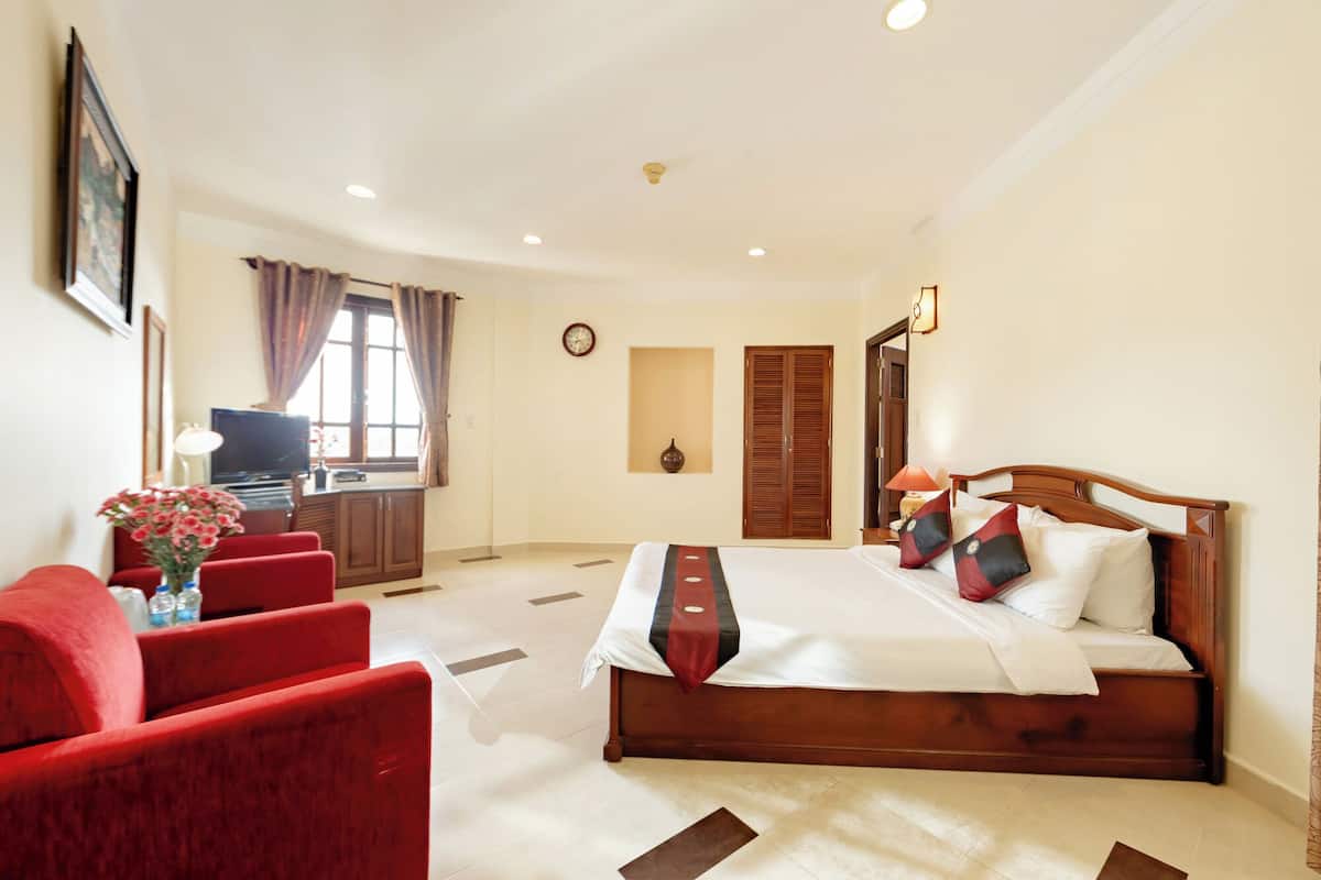 A hotel room with a bed, red armchairs, a desk with a computer, and a window with curtains.